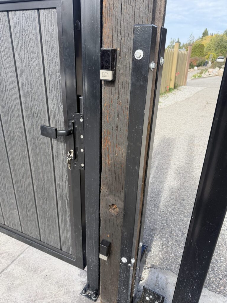 Photo eyes mounted on a gate post beside a black composite cantilever gate, with a DEA gate operator at its base